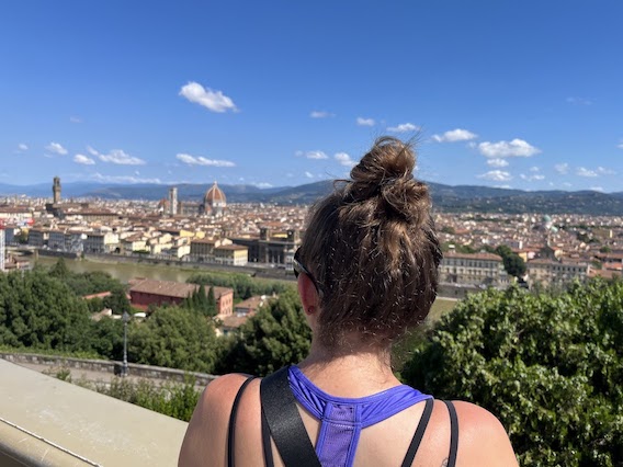 A woman with brown hair and a purple tank top, looking out over Florence, Italy. The picture is taken from behind so you can't see her face.